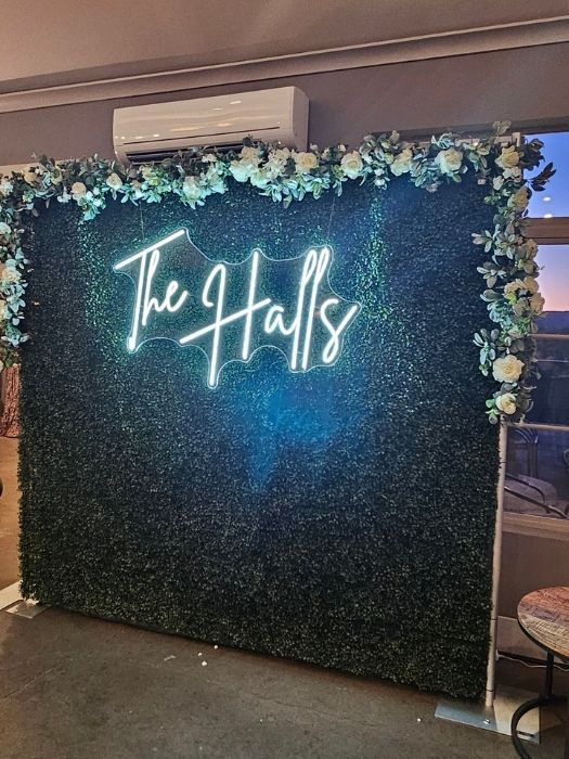 Boxwood greenery wall backdrop with floral trim and custom neon 'The Halls' sign, designed and rented by Simply LaMayed Custom Decor.
