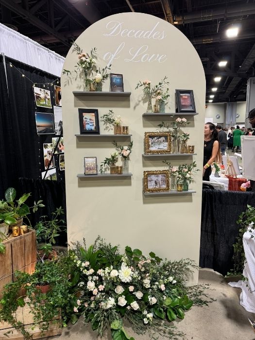 Arched photo display wall with 'Decades of Love' design, decorated with floral greenery and gold picture frames, available for event rentals from Simply LaMayed.