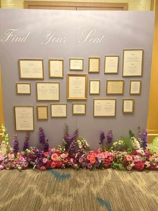 Elegant seating chart wall with gold-framed table assignments and a lush floral base, created by Simply LaMayed for weddings and formal events.